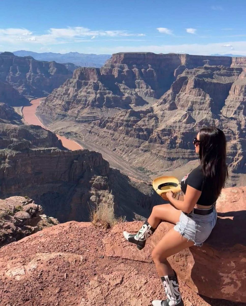 Traveller sitting on the edge of the Grand Canyon, wearing cowboy boots and holding a hat and looking out over the Colorado River and layered red cliffs.