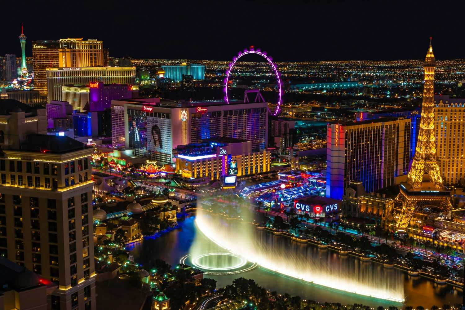 Night view of the Las Vegas Strip with the Bellagio fountains, Paris Las Vegas Eiffel Tower, and High Roller observation wheel illuminated in neon lights.